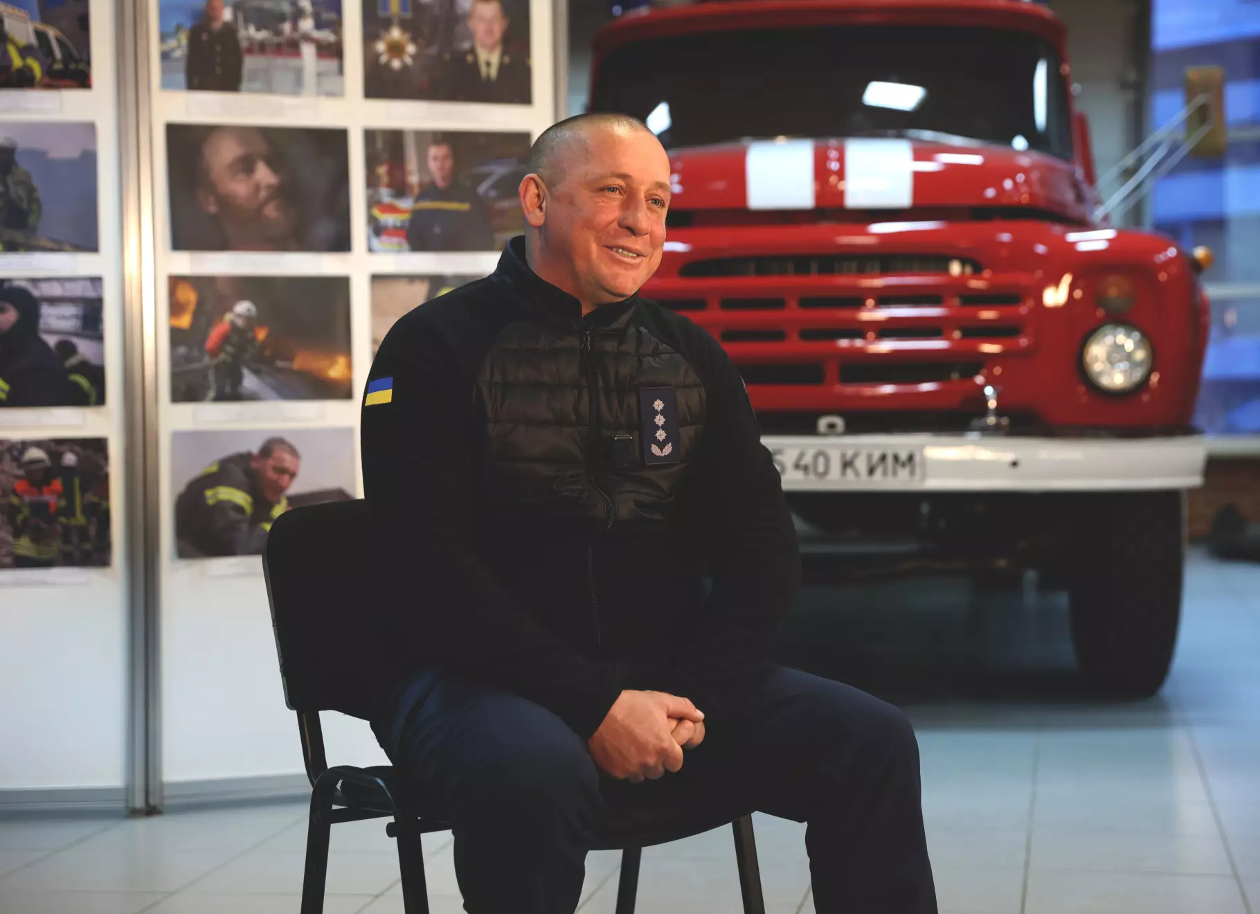 Чоловік у формі ДСНС сидить на стільці, на фоні червона пожежна машина та стіна з фотографіями
