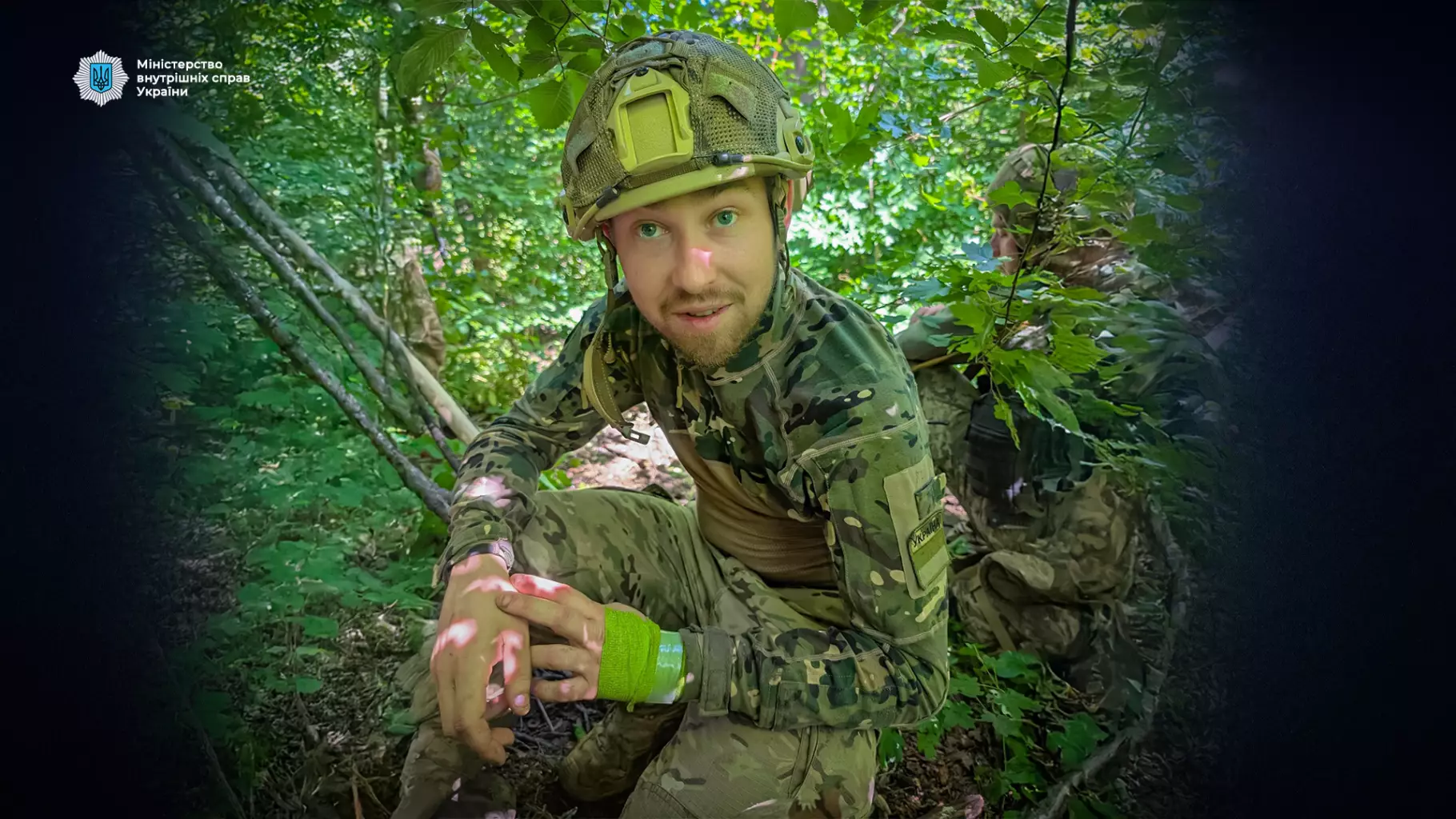 Прикордонник у камуфляжі сидить у лісі, тримаючи руки на колінах, поруч інший військовий серед зелені