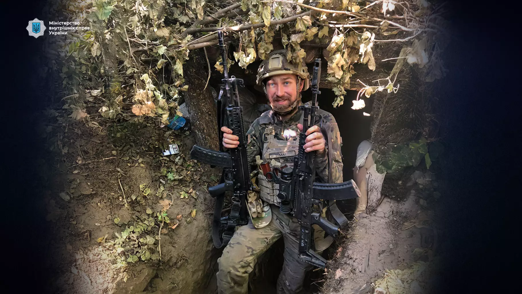 Український прикордонник у камуфляжі з автоматами, сидить у окопі, оточений листям та гілками