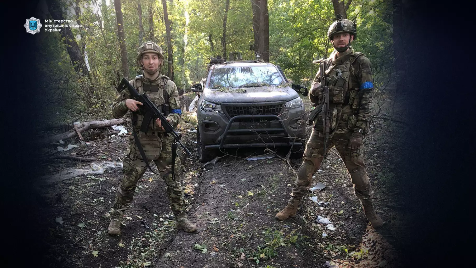 Двоє українських прикордонників у камуфляжі стоять перед броньованим автомобілем у лісі