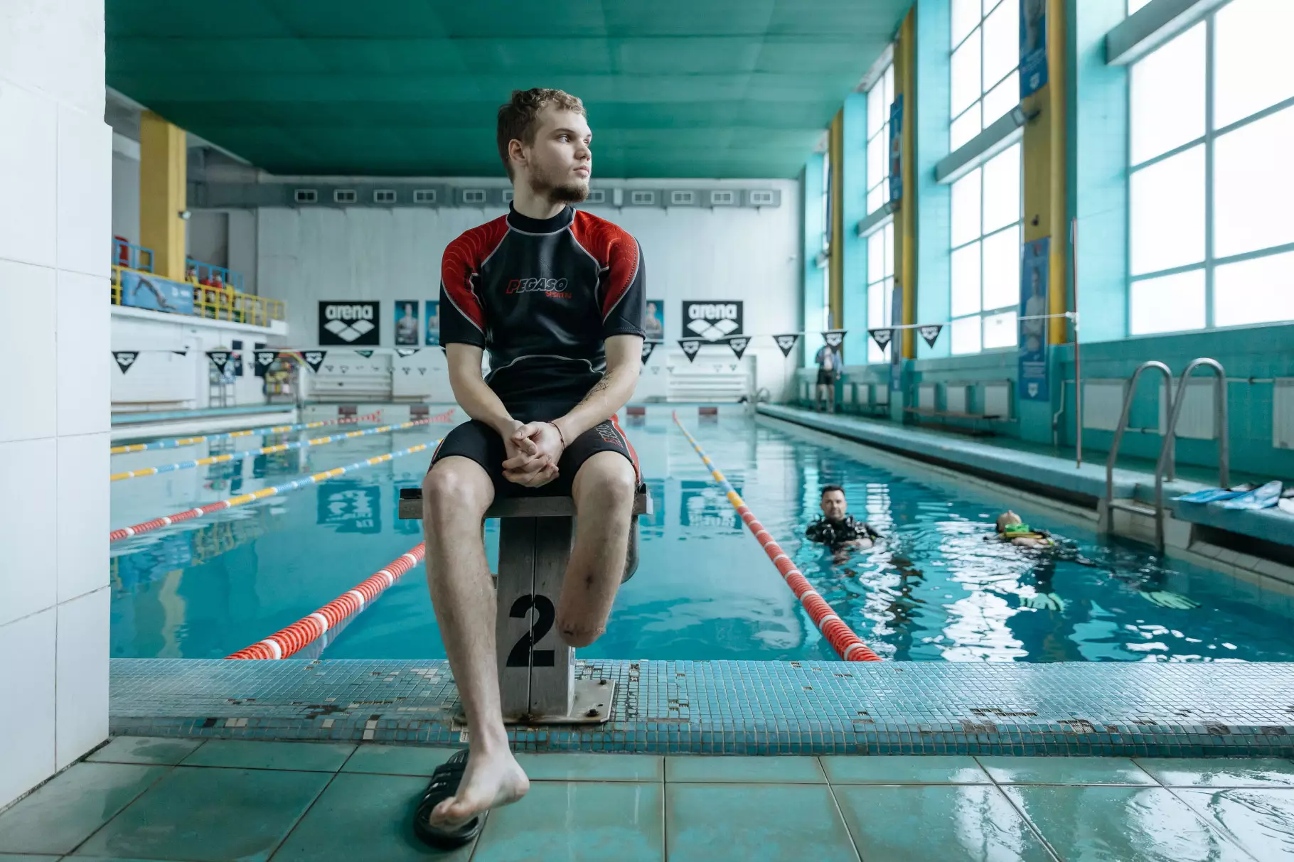 Чоловік у спортивному одязі сидить на стартовій тумбі біля басейну, де тренуються дайвери