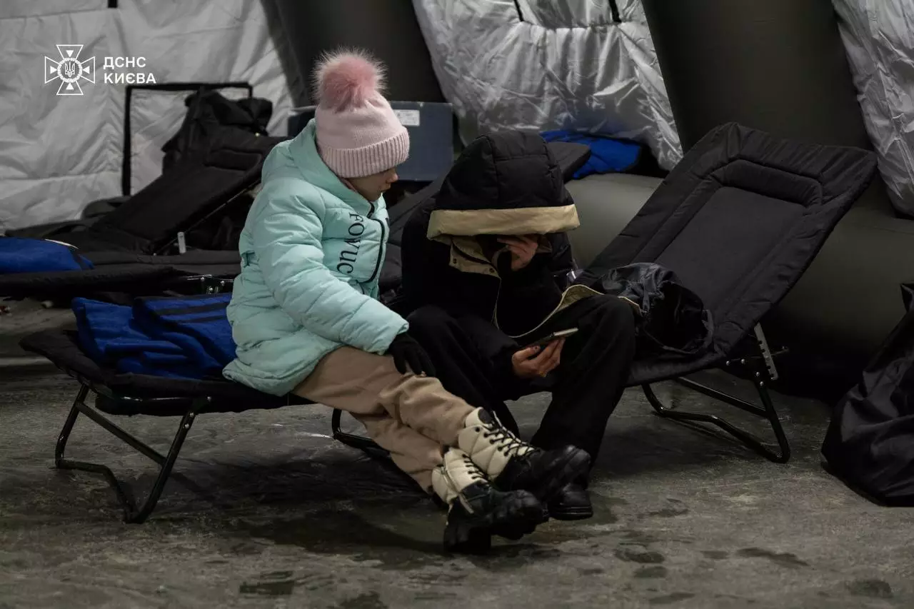 Дитина та дорослий сидять на розкладних ліжках у тимчасовому притулку після атаки на інфраструктуру