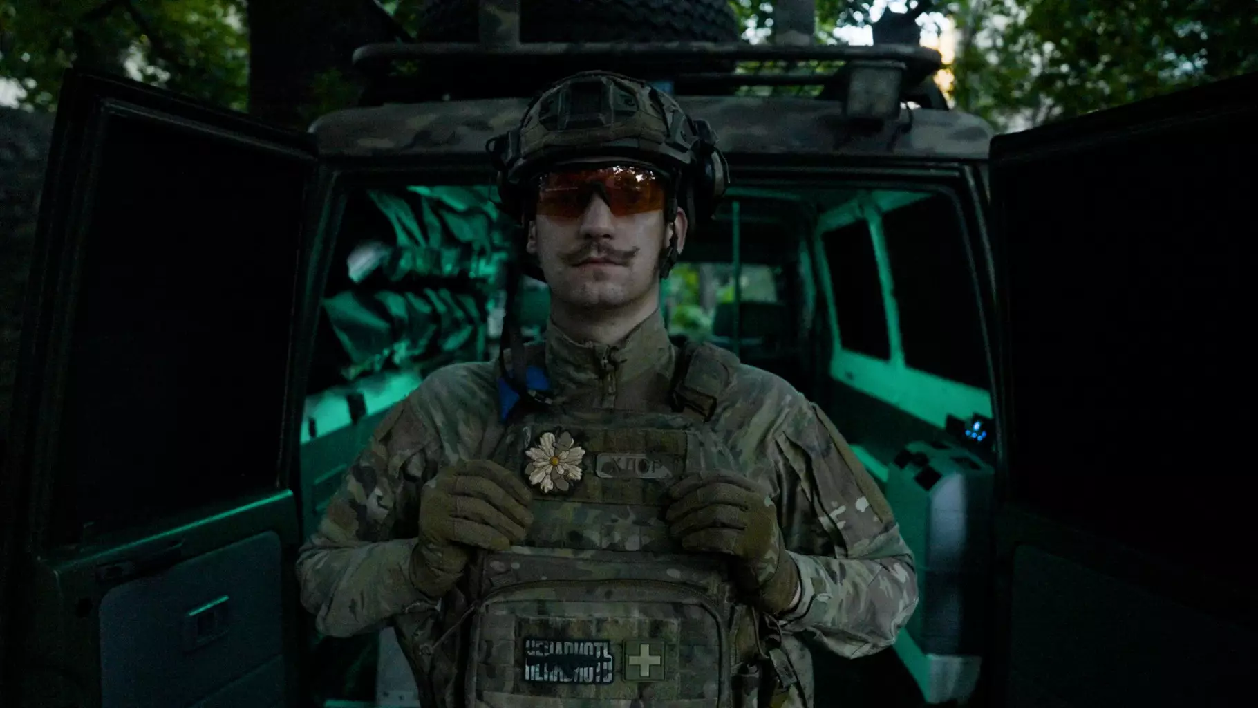 Солдат у військовій формі стоїть перед військовим транспортом, тримаючи медичний рюкзак