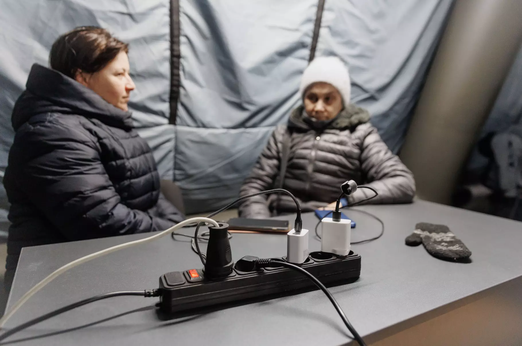 Дві людини в теплому одязі сидять у наметі, заряджаючи пристрої від подовжувача