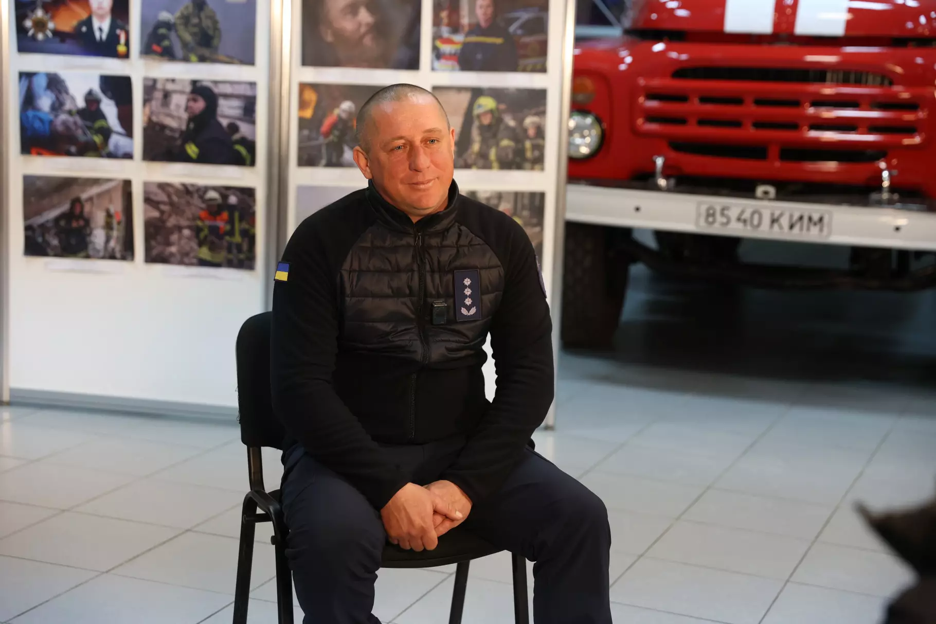 Рятувальник ДСНС України сидить на стільці в приміщенні з фотографіями та червоним пожежним авто на фоні
