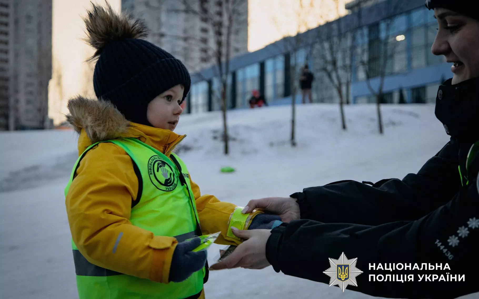 Дитина в жовтому жилеті отримує світловідбивальний елемент від поліцейського на засніженій вулиці