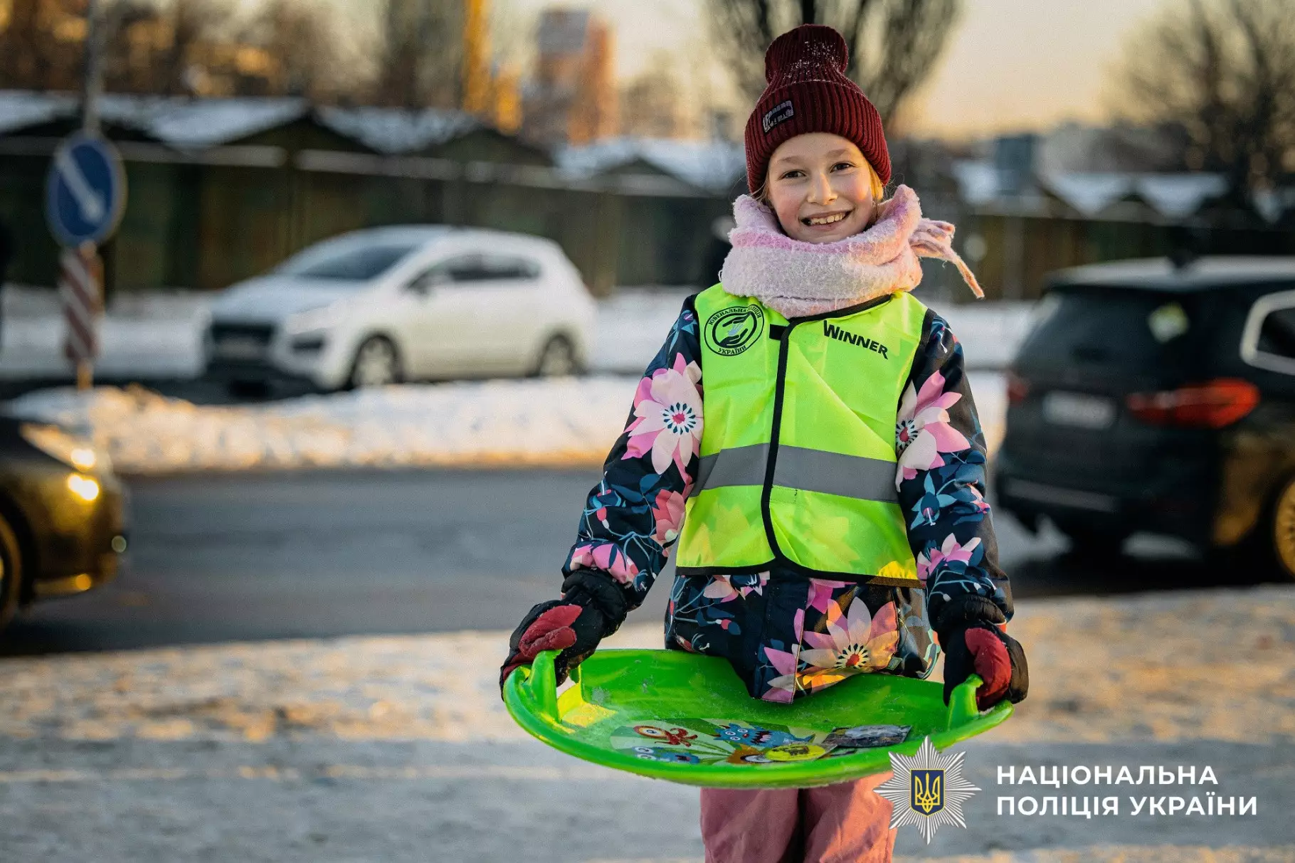 Дитина в світловідбивальному жилеті тримає санки на засніженій вулиці, поруч автомобілі