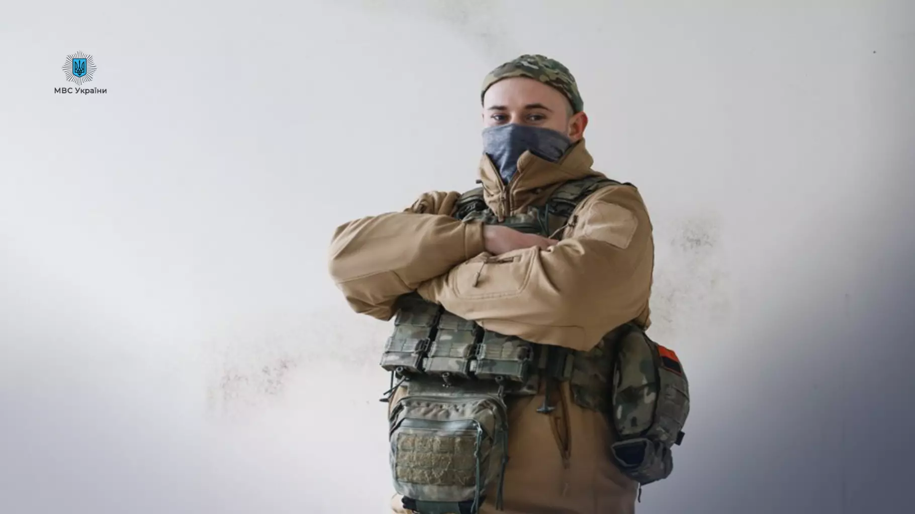 Прикордонник у військовій формі з маскою на обличчі стоїть зі схрещеними руками на фоні стіни