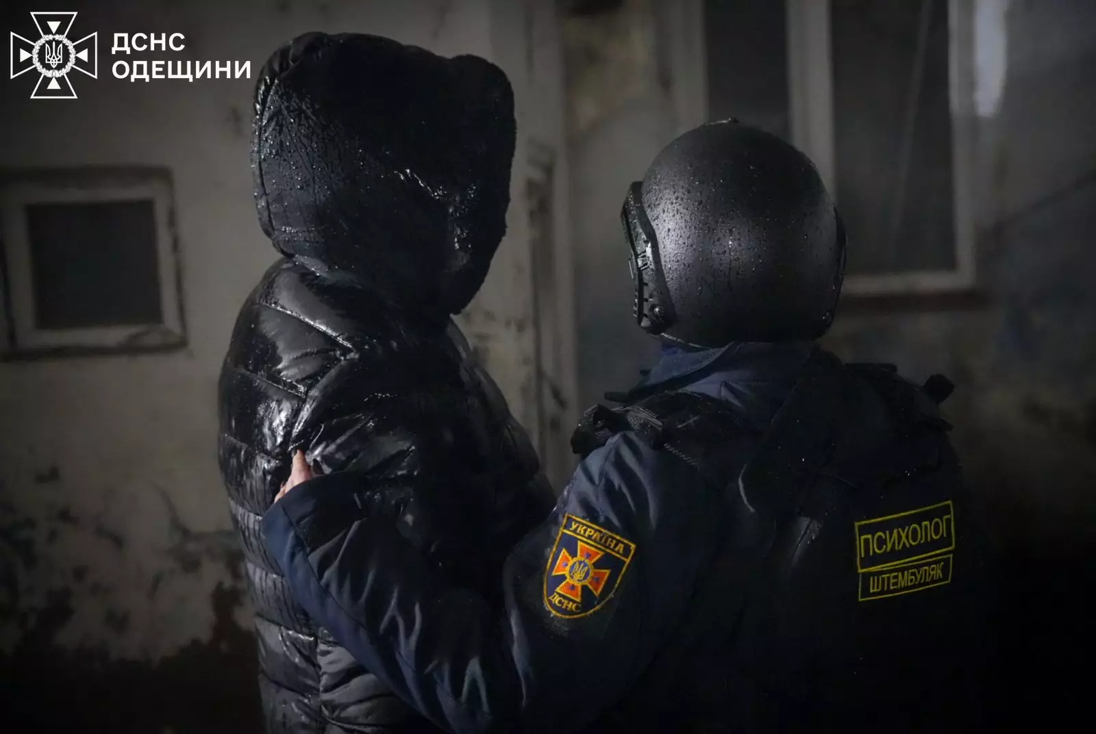 Поліцейський у шоломі підтримує людину в чорній куртці на тлі пошкодженої будівлі