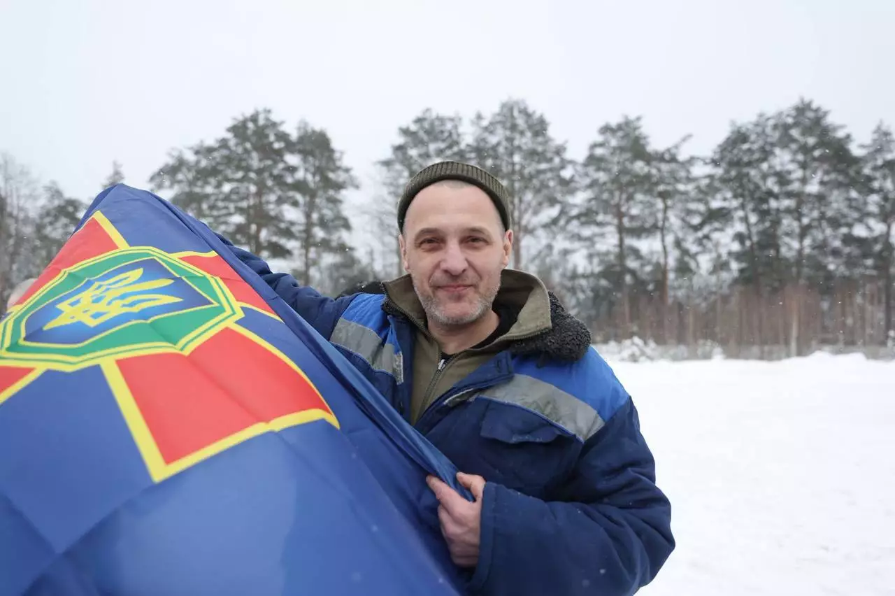 Чоловік у зимовому одязі тримає прапор з емблемою, на фоні засніжений ліс