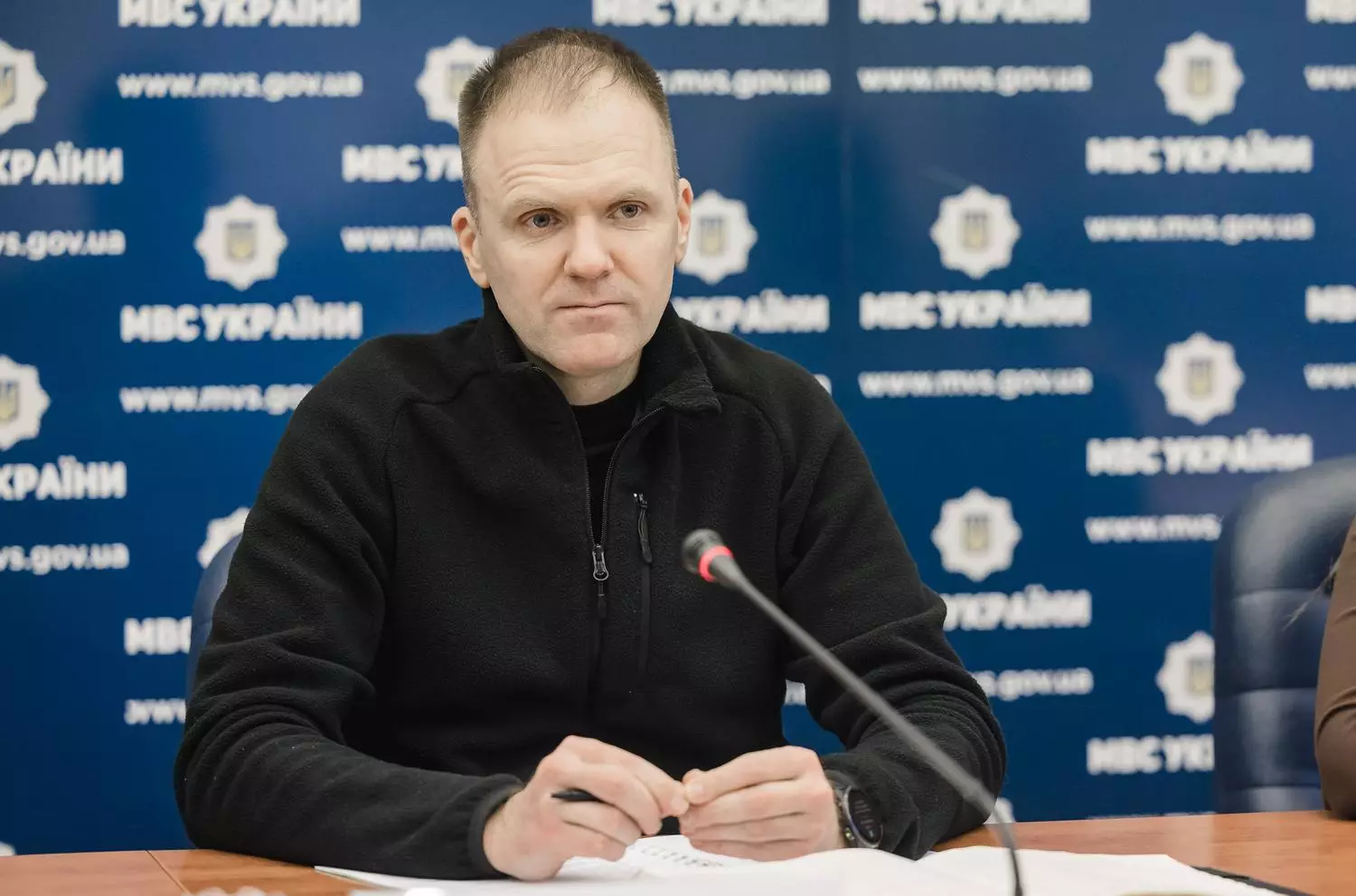 Чоловік у чорному светрі сидить за столом на офіційній зустрічі, перед ним мікрофон і документи