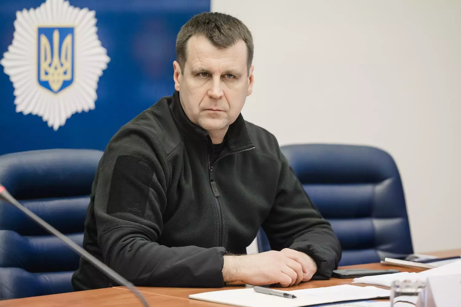 Чоловік у чорній куртці сидить за столом на офіційній зустрічі, на задньому плані герб України