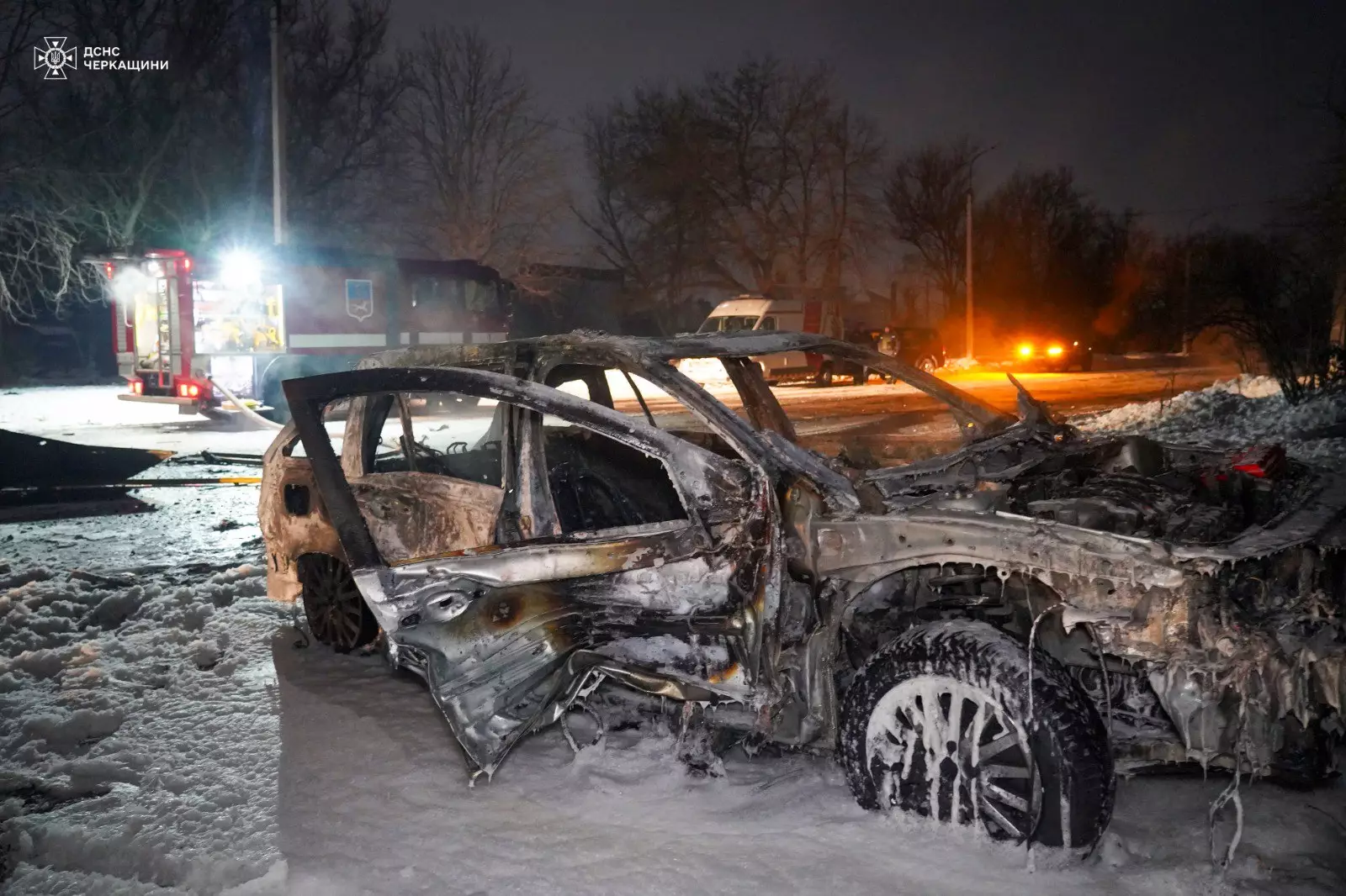 Згорілий автомобіль на засніженій вулиці, поруч працюють рятувальники, видно пожежну машину