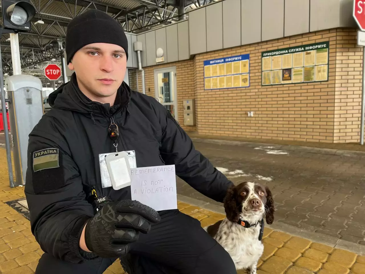 Прикордонник з собакою тримає табличку з написом "REMEMBRANCE IS NOT A VIOLATION" на тлі будівлі