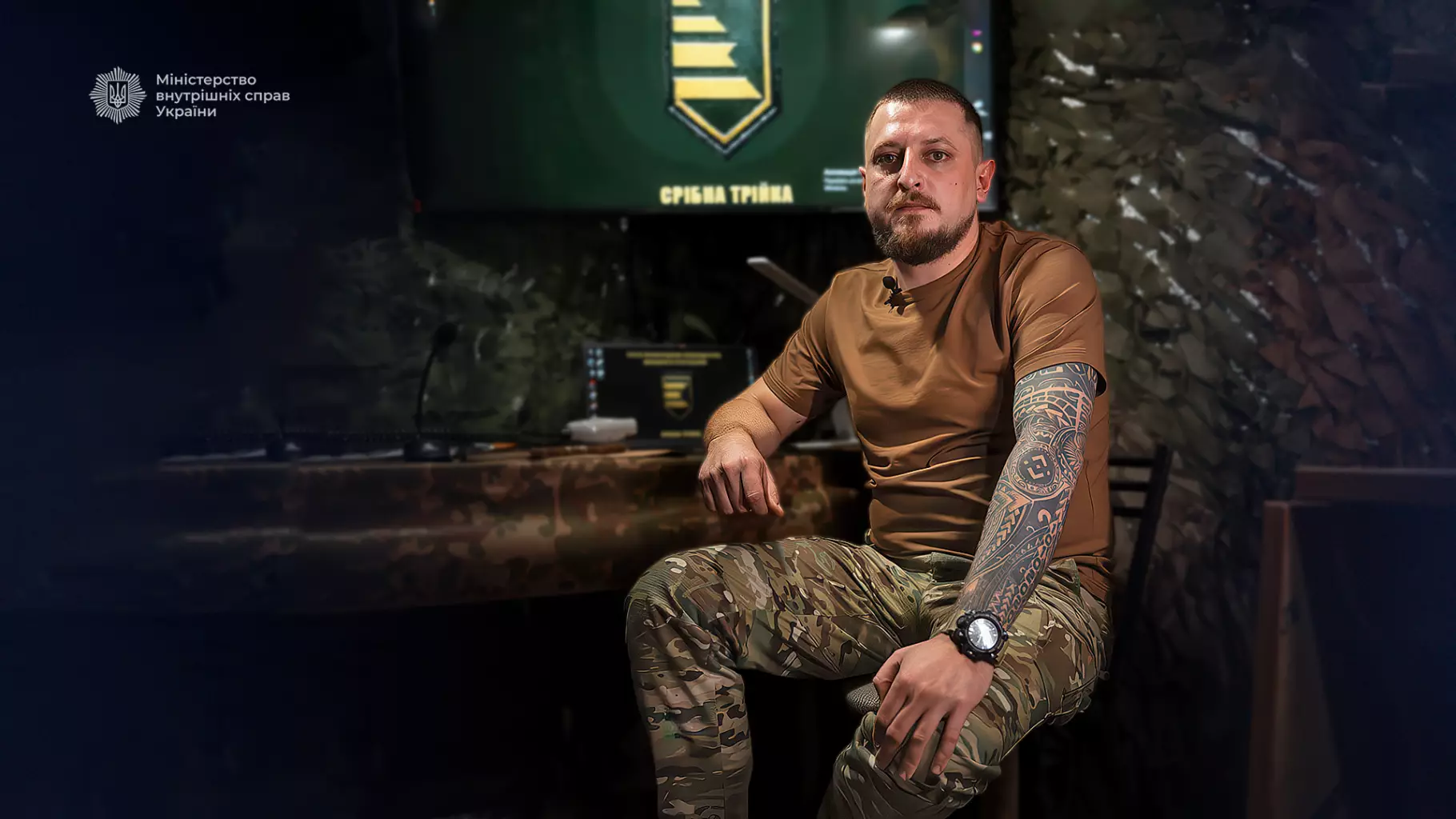 Чоловік у військовій формі сидить у кімнаті з камуфляжним декором, на задньому плані емблема служби