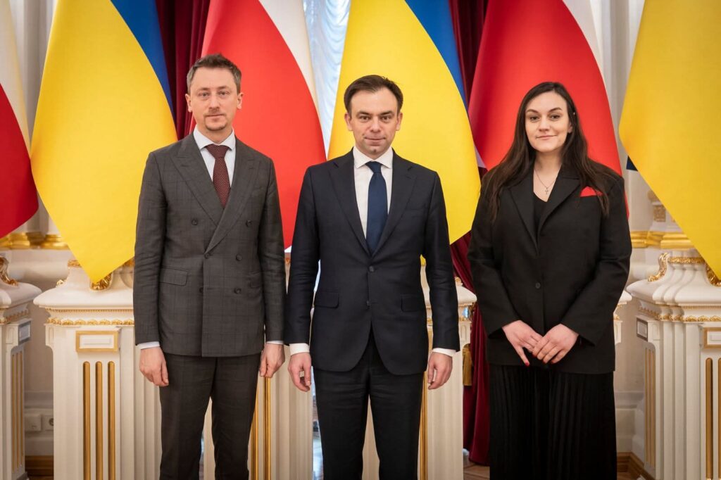 Три офіційні особи стоять перед прапорами України та Польщі під час дипломатичної зустрічі