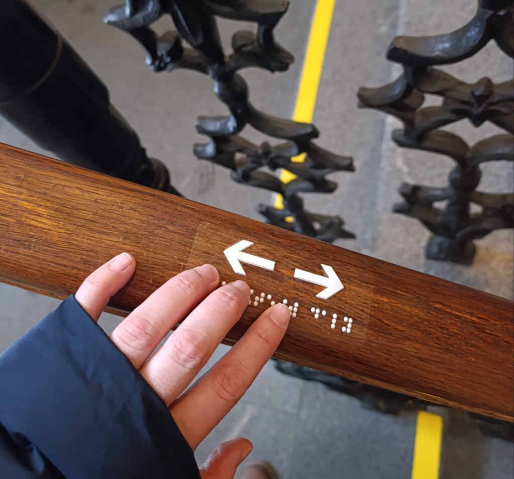 Рука торкається тактильного вказівника зі шрифтом Брайля на дерев'яних перилах у філармонії