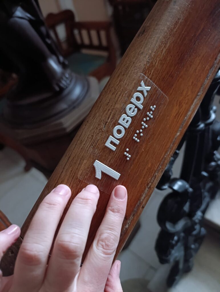 Рука торкається тактильного вказівника з шрифтом Брайля на перилах у Національній філармонії України