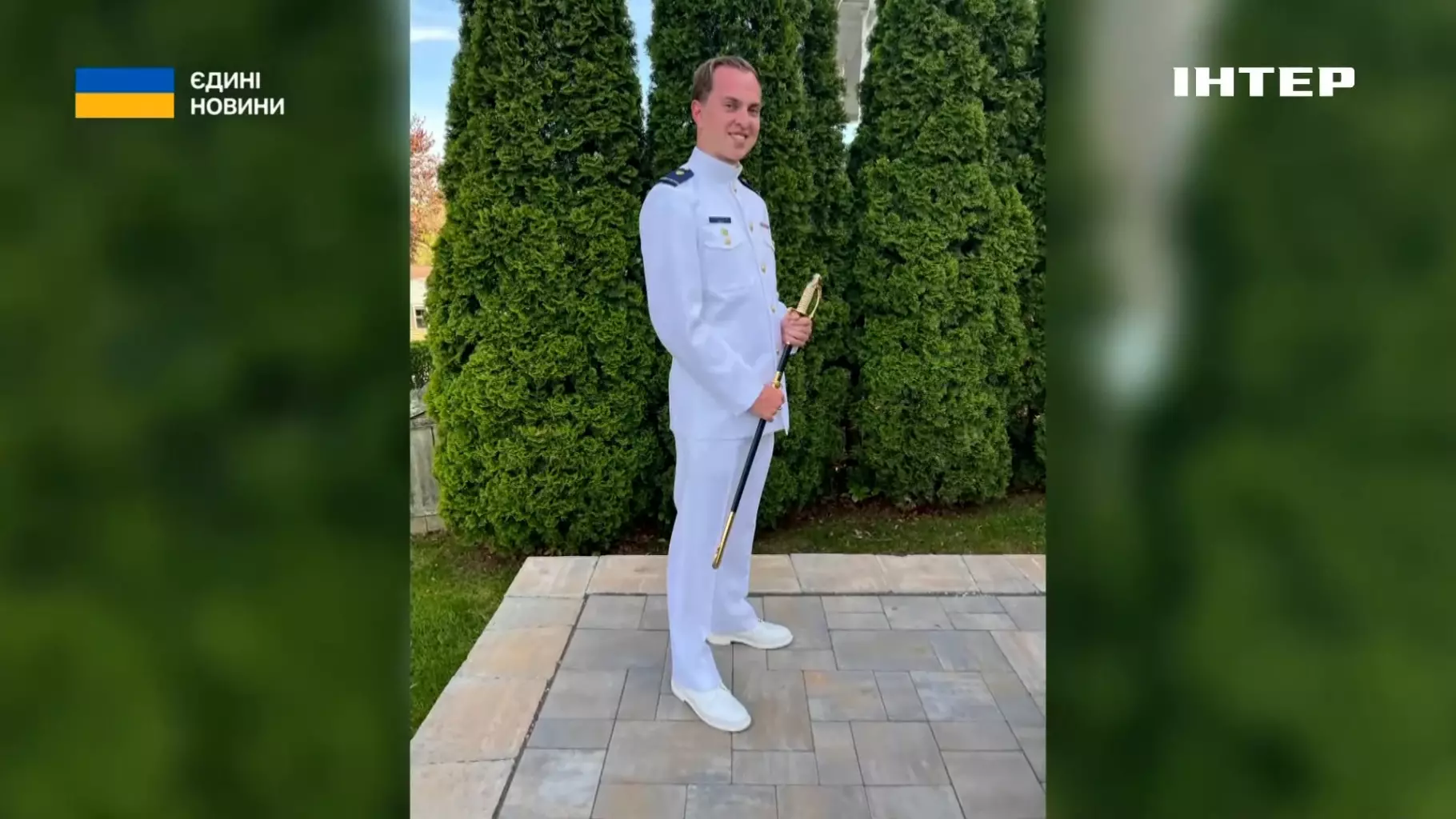 Офіцер морської охорони у парадній формі стоїть на тлі зелених дерев, тримаючи шаблю