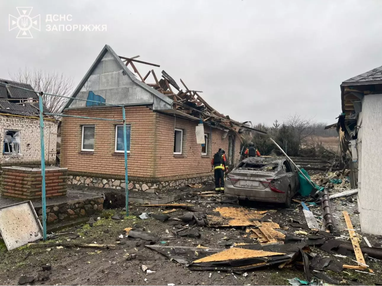 Рятувальники оглядають зруйнований будинок після обстрілу, пошкоджене авто на передньому плані